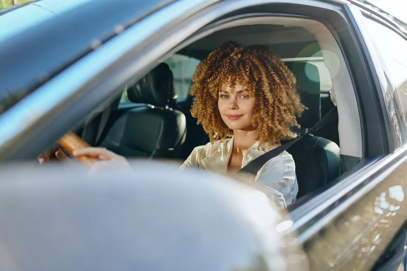 Smiling woman driving car with seatbelt fastened, curly hair and casual clothing, bright daylight and modern vehicle interior visible.
