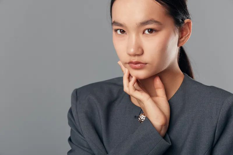 Young woman with clear skin and dark hair wearing a gray blazer, posing with hand near face against gray background, showing natural beauty and calm confident expression.