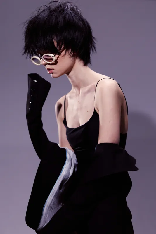 Stylish woman with short black hair wearing round glasses and a sleeveless black outfit in a minimalistic setting