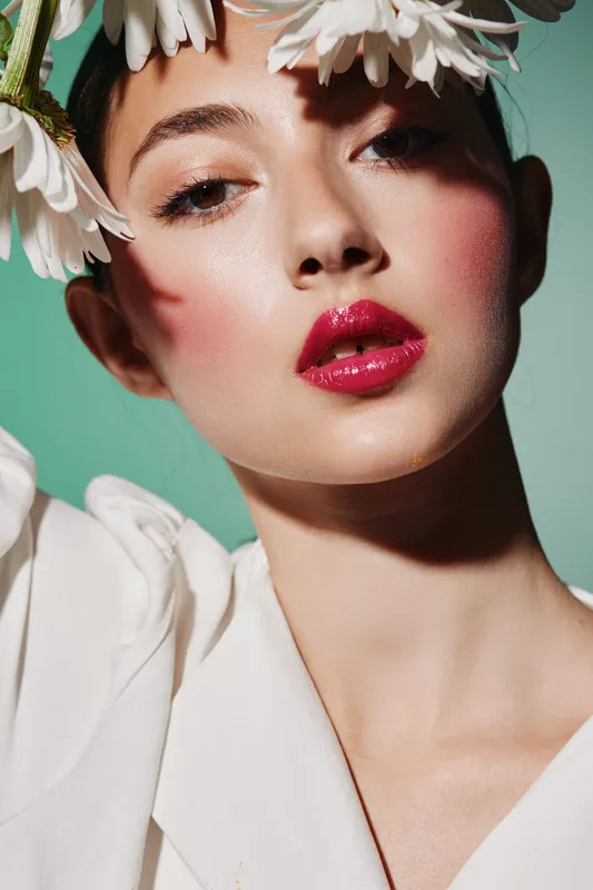 Close-up portrait of a beauty model with glossy red lips, soft pink blush, and natural eyebrows wearing large white flowers on her head on a green background.
