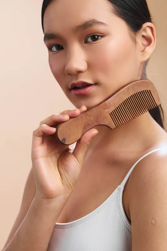 asian woman gently holds a wooden comb near her hair in a clean studio setting, exuding calm self care and natural beauty through soft lighting and a minimal aesthetic.