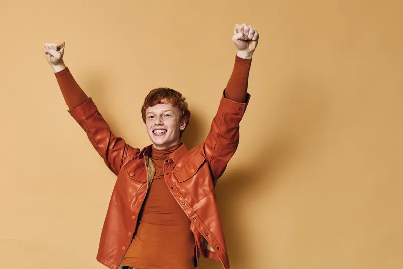 Young happy man with red hair wearing brown jacket and sweater raising arms up in celebration smiling with joy. Isolated background with positive emotion and success gesture.