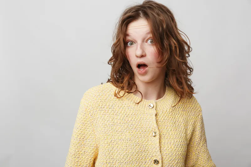 Surprised young woman with curly hair wearing yellow knitted sweater looks shocked with open mouth on grey background. Concept of unexpected news, emotion, and human reaction.