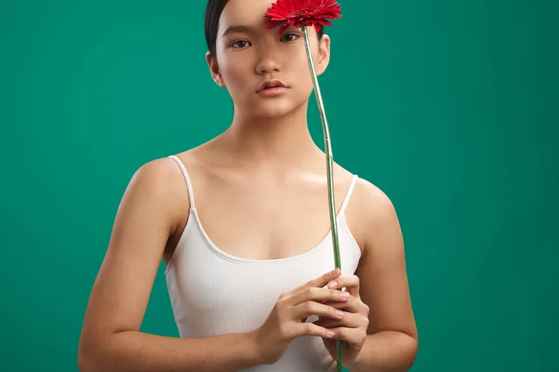 asian woman in a white tank top stands against a teal backdrop, holding a red flower with both hands, calm expression, soft studio lighting, minimal portrait