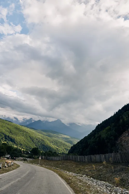 Road winds through a mountainous valley under a cloudy sky, green slopes and distant peaks creating a tranquil expansive nature scene for travel and outdoor photography.