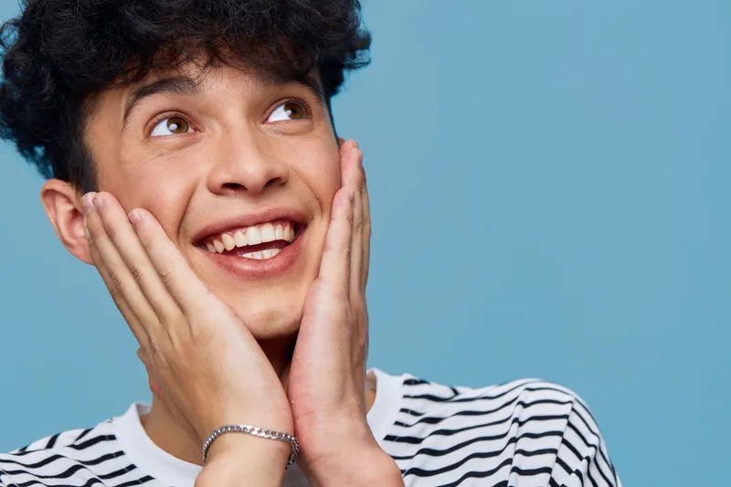 Smiling young man with curly hair wearing striped shirt, expressing joy and surprise against a light blue background, casual style and bright mood.