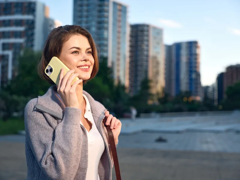 A cheerful businesswoman speaks on a smartphone outdoors, smiling as she chats near modern high-rise buildings. The urban setting conveys confidence, optimism, and professional energy.