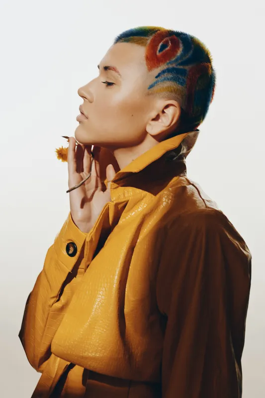 fashion woman with colorful shaved hair and artistic patterns wearing a yellow leather jacket in a studio with soft lighting and a neutral background