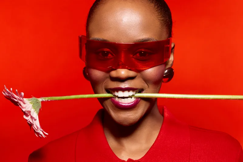 Smiling African woman with dark skin wearing red sunglasses and red clothing holds a pink flower in her mouth against a bright red background. Concept for fashion, beauty, and confidence.