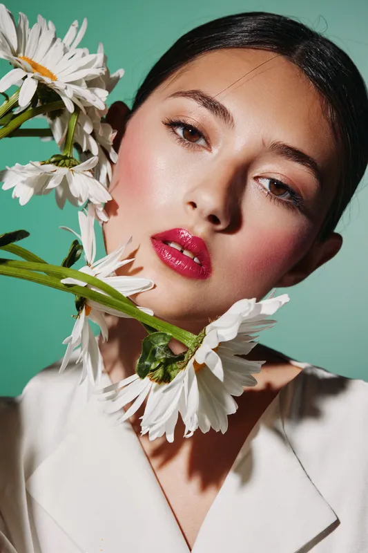 Young woman with glossy red lips and dewy skin poses holding white daisies. Soft lighting highlights her fresh makeup and smooth complexion. Concept of beauty, fashion, and creative portrait.