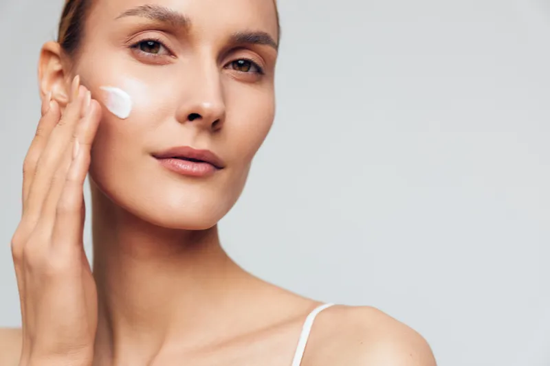 Young woman applying face cream on cheek, skincare routine, natural beauty, smooth skin, soft lighting, closeup portrait, neutral background, healthy complexion