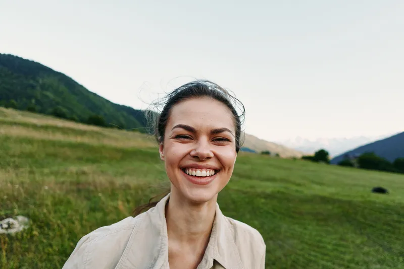 A smiling woman in an outdoor field stands with a bright landscape and distant mountains, capturing a natural, healthy lifestyle moment in daylight and nature backdrop.