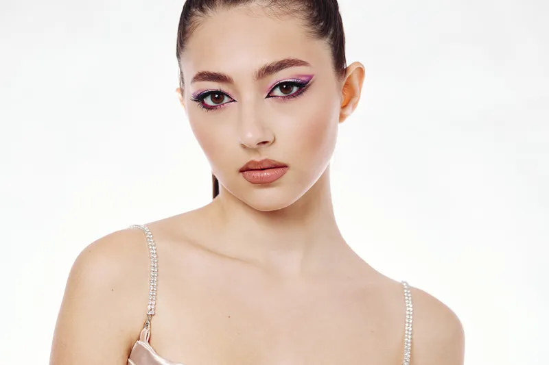 Young woman with creative pink eye makeup looks confidently at camera. She wears a strappy top with rhinestones. Studio shot on white background. Concept of beauty, fashion, and trend.