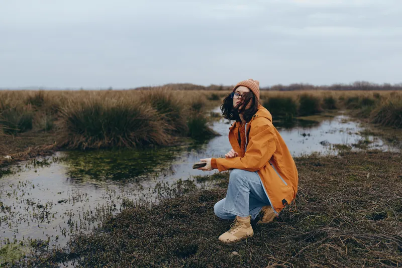 Woman kneeling outdoors in a wetland landscape, wearing a jacket and boots, calm lifestyle pose by shallow water and grass, overcast winter sky, peaceful nature mood, soft light on ground