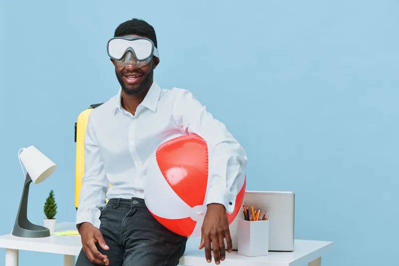 Casual smiling man with dark skin holding a red and white beach ball, wearing white shirt and black pants, in a bright modern workspace with blue background, summer and leisure concept.