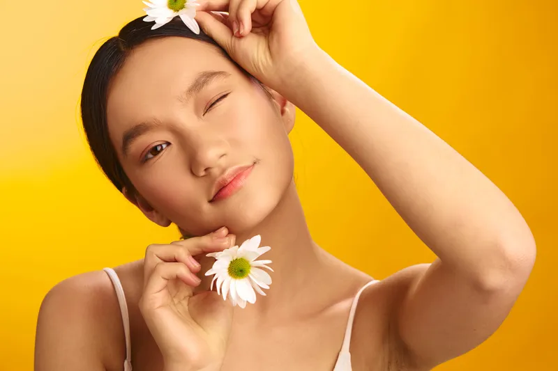asian woman holds a daisy near her cheek, smiling softly against a vivid yellow backdrop, conveying fresh beauty, optimism, and a cheerful mood.
