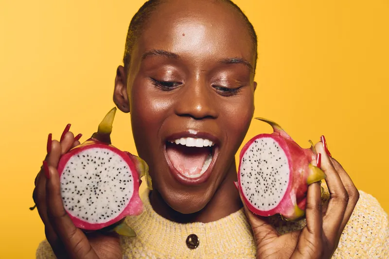 Happy African woman with dark skin holds two halves of ripe dragon fruit near face, smiling with open mouth on yellow background. Concept of healthy lifestyle, exotic fruit, and joy.