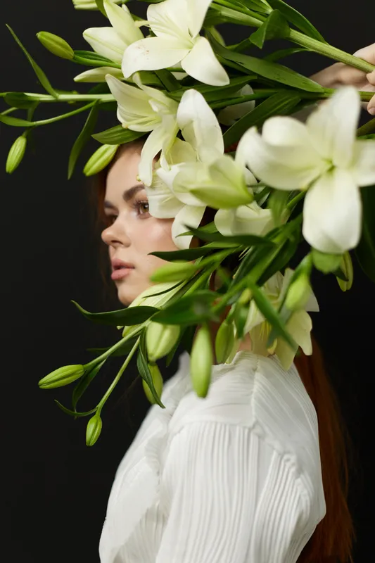 Elegant young woman with long brown hair adorned with white lilies, showcasing a striking floral crown, exuding serenity and beauty against a dark background