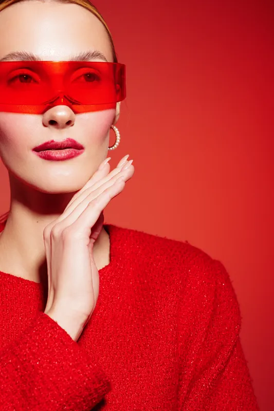 Stylish woman in red sweater and red transparent glasses poses with hand near face on red background. Concept of fashion, beauty, modern style, and confident female portrait.