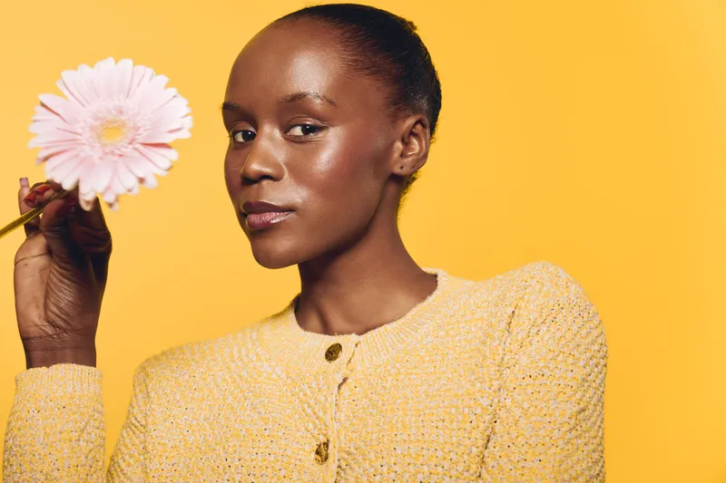 Portrait of a confident black woman with dark skin holding a pink flower against a yellow background. Concept of beauty, natural skincare, and self-confidence in studio lighting.