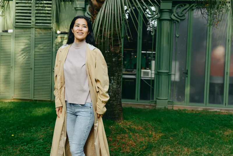 Woman wearing a beige trench coat and light sweater stands outdoors on a lush lawn near glass buildings, showcasing casual fashion style, relaxed pose, natural light, and urban garden ambiance