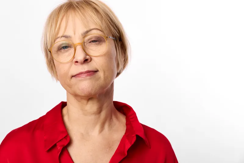 Senior woman portrait, close face view, glasses, red shirt, studio lighting on white background, calm expression, neutral posture, looking at camera, soft natural tones