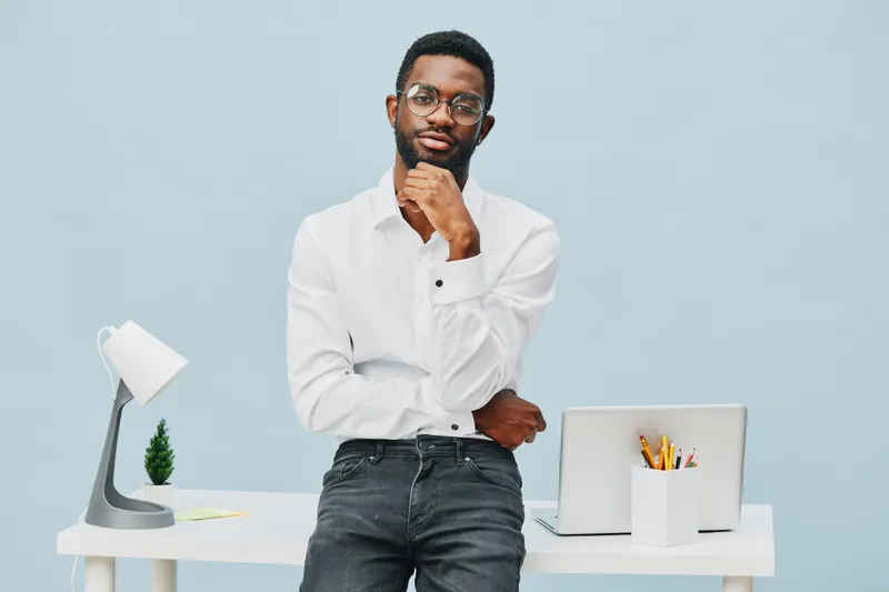 Confident young African American man in white shirt standing with hand on chin at modern office workspace. Simple desk with laptop, lamp, stationery. Inspiration, professionalism, smart casual style c