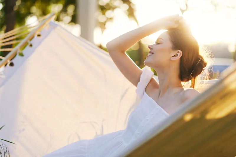 Young woman enjoying a sunny afternoon in a hammock, wearing a light dress, radiating happiness among natural greenery, capturing a serene summer vibe