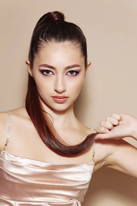 Young woman with perfect makeup wearing a satin dress holds her shiny ponytail, captured on beige background. Concept for beauty, creative, fashion, elegance.