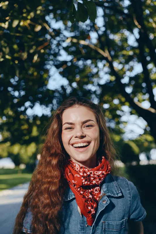 lifestyle woman with long curly hair wearing modern boho-western denim street style and a red bandana, smiling outdoors in natural sunlight with film color effect