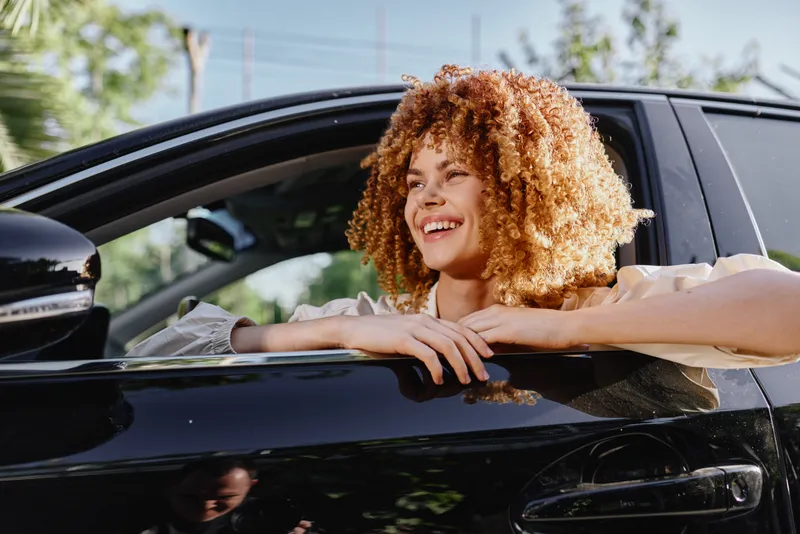smiling woman with curly hair leaning out of black car window, natural light, outdoor, joyful expression, casual clothing, sunny day
