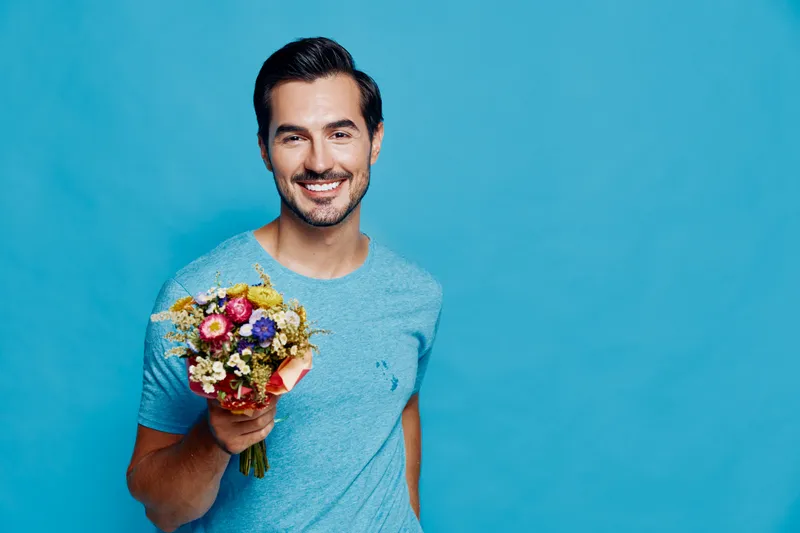 Smiling man in blue t-shirt holding bouquet of flowers, looking cheerful while posing against vibrant blue background. Floral expression of happiness concept.