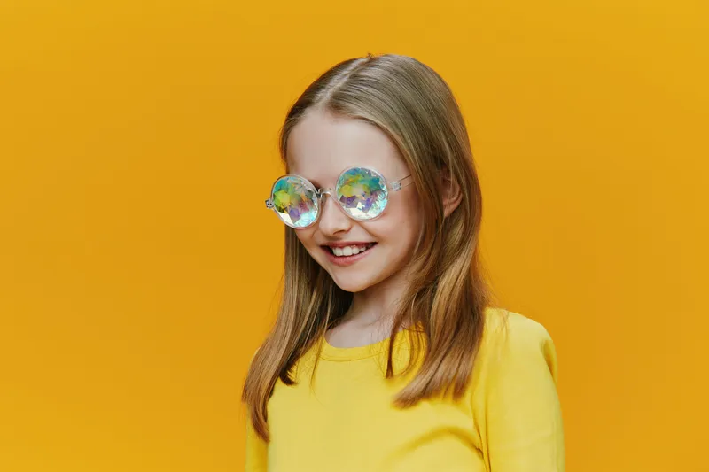 Cheerful girl wearing colorful glasses, smiling against a bright yellow background, perfect for children s fashion or summer themes