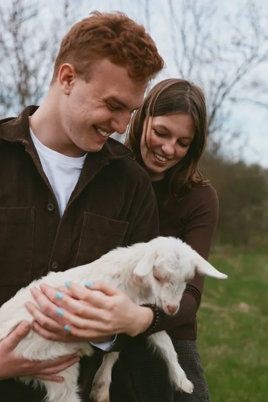 A joyful couple outdoors with a playful goat kid, smiling as they cradle the young animal. A lifestyle scene about companionship, caregiving, and rustic life in the countryside.