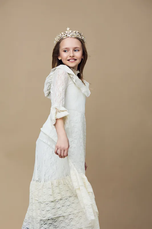 Young girl wearing a delicate white dress and crown, smiling gracefully against a soft beige background, exuding joy and innocence, perfect for children s fashion