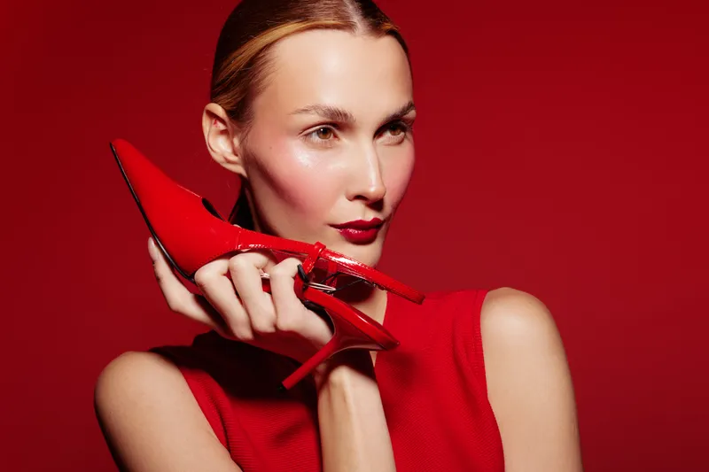 Stylish woman in red sleeveless top holds red high heel shoe near face on red background. Concept of fashion, elegance, beauty, and bold color contrast in studio lighting.