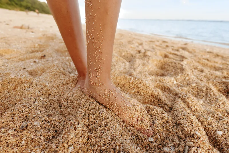 Close-up of bare feet standing on sandy beach with grains of sand falling down. Concept of summer vacation, relaxation, and natural outdoor environment by the sea.