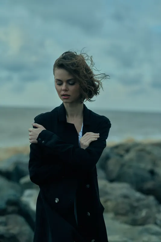 Woman in a dark coat stands at the windy sea edge with crossed arms, moody mood, ocean backdrop, stormy sky, reflective pose, fashion scene, solitary moment