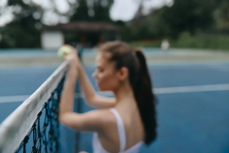 Young woman with long hair in a sporty outfit, standing by the tennis net, expressing focus and determination on a blue tennis court