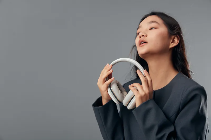Young woman holding headphones near face with eyes closed, enjoying music, expression of relaxation and calm in studio with neutral gray background and casual dark clothing.