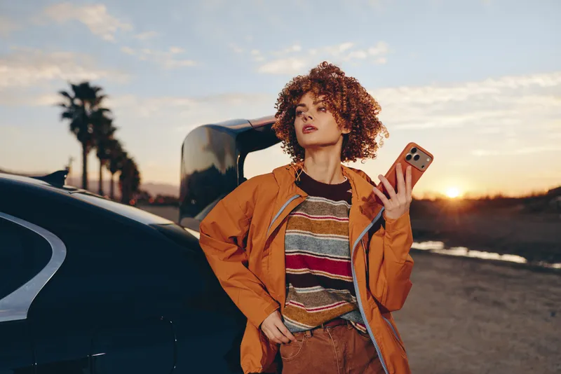 Cheerful woman with curly hair outdoors by a car at sunset, holding a smartphone, relaxed lifestyle style, orange jacket, warm evening light, quiet road background soft breeze and city sky