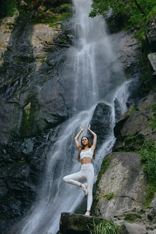 woman practicing yoga outdoors in white sportswear stands in tree pose near a large waterfall surrounded by rocks and lush green foliage