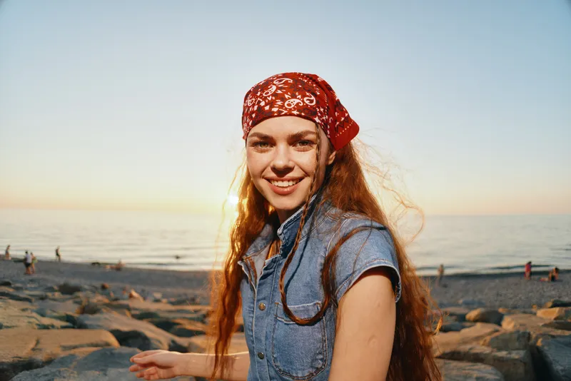 Lifestyle woman wearing modern boho-western denim street style with a red bandana, smiling by the beach at sunset in warm film color tones and natural light.