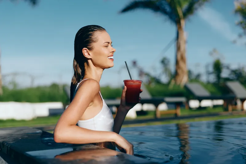 Smiling woman in a white swimsuit leans on a pool edge, holding a refreshing drink. Sunny poolside scene conveys leisure, wellness, and summer relaxation.
