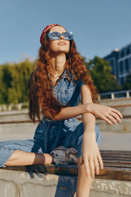 lifestyle woman in modern boho-western denim street style with long curly hair wearing sunglasses and a red bandana enjoying sunny day urban vibe film color