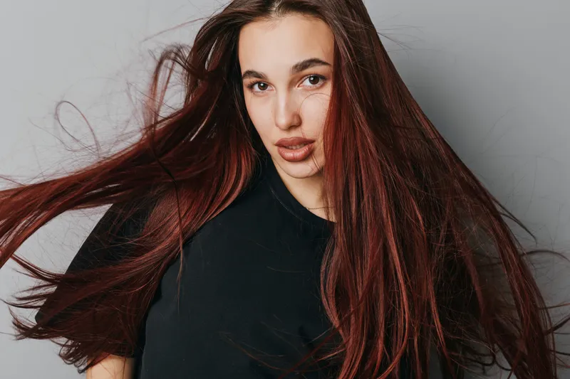 Young woman with long flowing hair and natural makeup posing in black shirt against neutral background. Portrait of confident female with loose hair and serious expression.