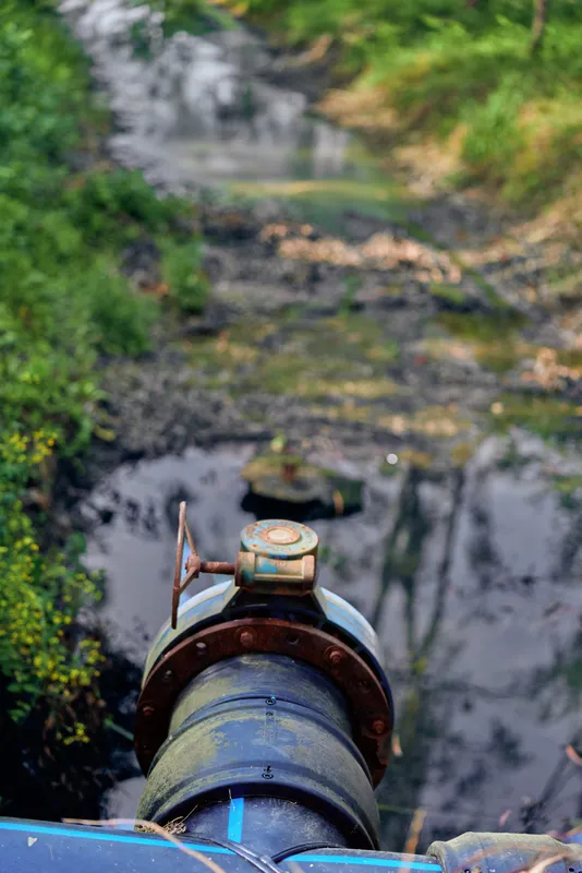 Pipe valve with rusted metal and flowing water over a muddy stream outdoors, closeup of industrial connection, corrosion details and nature background for infrastructure inspection
