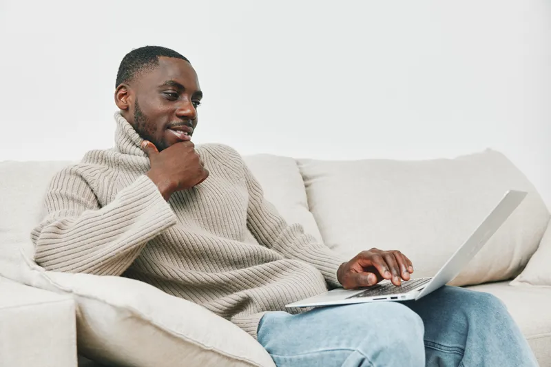 Young African man in cozy sweater using laptop at home, showcasing comfort and modern lifestyle, focusing on work and relaxation in a contemporary setting