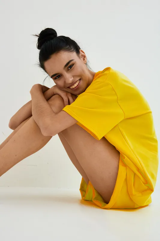 Smiling woman wearing bright yellow casual outfit sitting on white floor with relaxed posture and happy expression, presenting cheerful and vibrant mood.