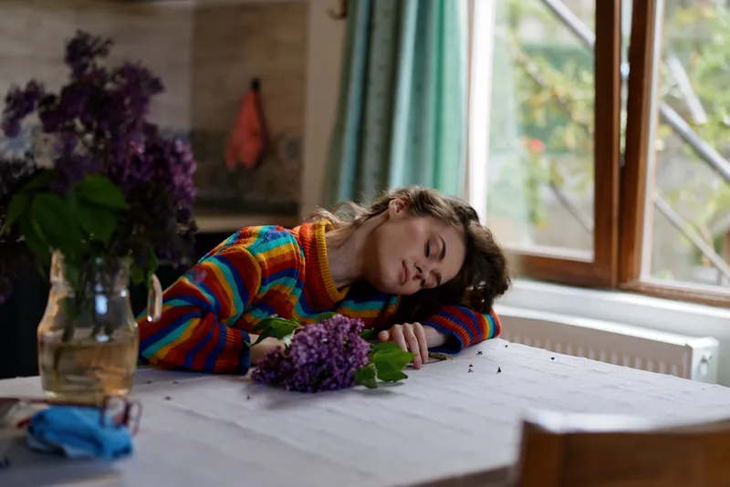 Young woman lifestyle at home, rests her head on a table in a colorful sweater, surrounded by flowers in a vase. A cozy indoor setting conveys peaceful relaxation, contemplation, and gentle fatigue.